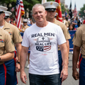 Man wearing RMRL Freedom Tee white shirt with Real Men Real Life and American flag graphic, marching alongside Marines at a patriotic parade