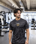 Tattooed man with curly hair in modern gym wearing RMRL Stone Grey Camo Dry-Fit Tee with Real Men Real Life center-chest logo
