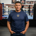 Hispanic man standing in boxing gym wearing RMRL True Navy Dry-Fit Tee with Real Men Real Life left-chest logo
