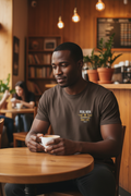 Man in warm coffee shop wearing dark chocolate RMRL Core Identity Collection tee with bold center-chest Real Men Real Life logo